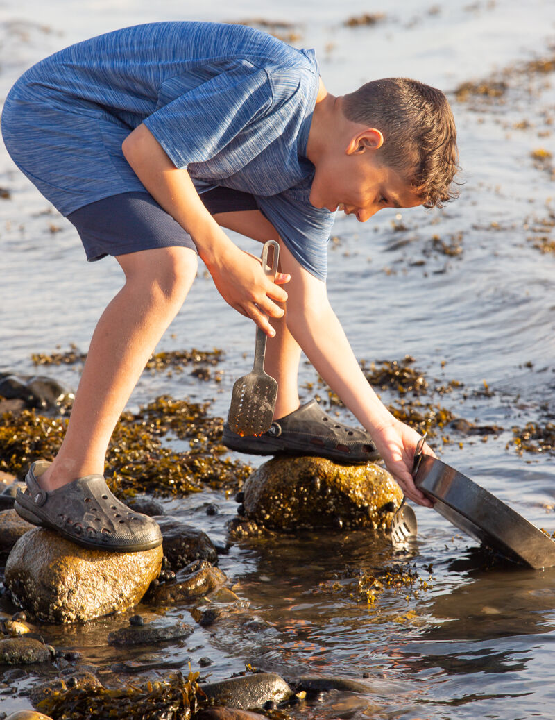 In the image, a young boy is exploring the water's edge. He is wearing a blue shirt, shorts, and water shoes. He is using a metal tool to examine something in the water. The boy appears focused and engaged in his activity, enjoying the outdoors.